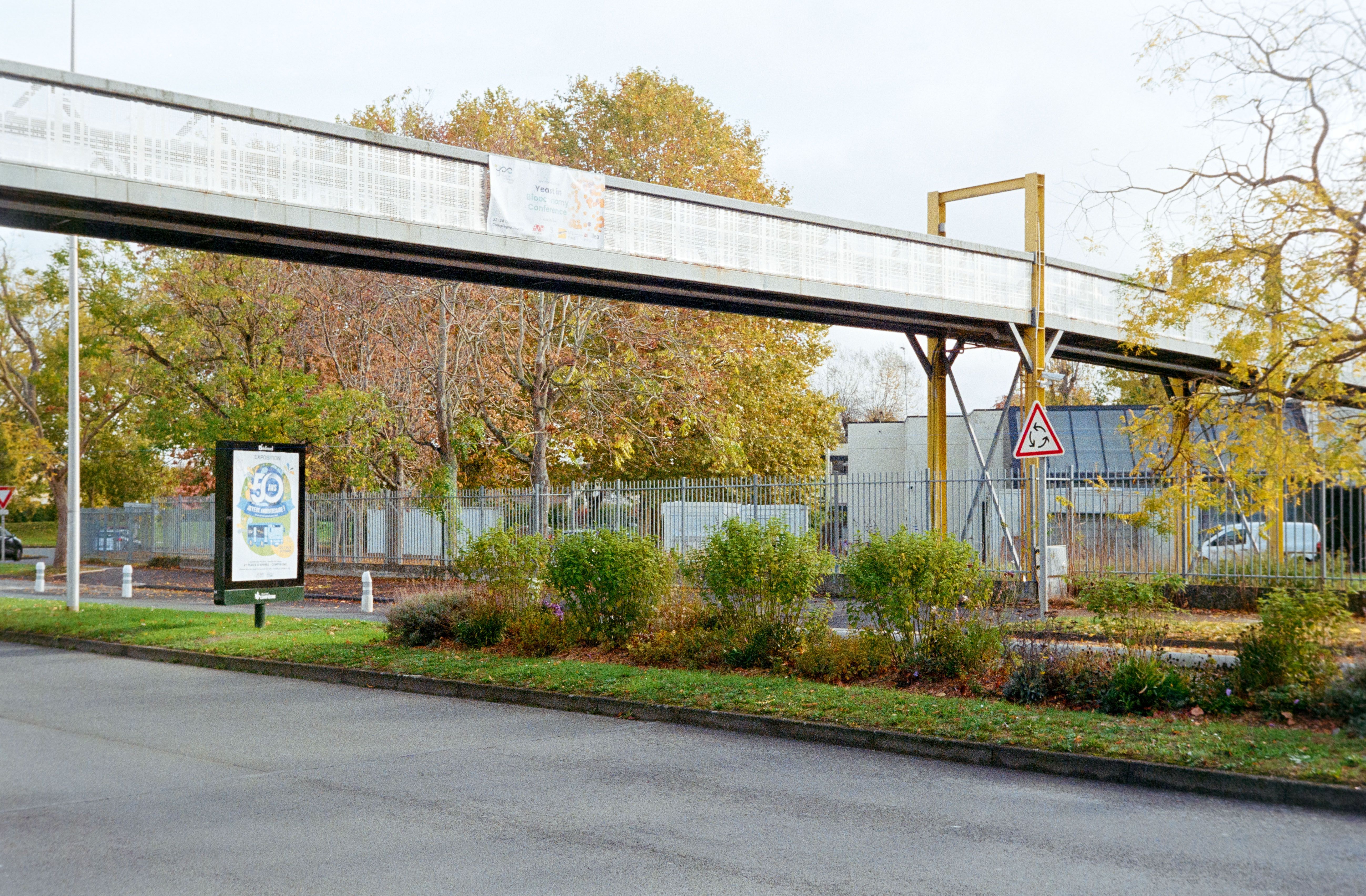 Elevated pedestrian bridge over a road in Compiègne, France, framed by vibrant autumn foliage.