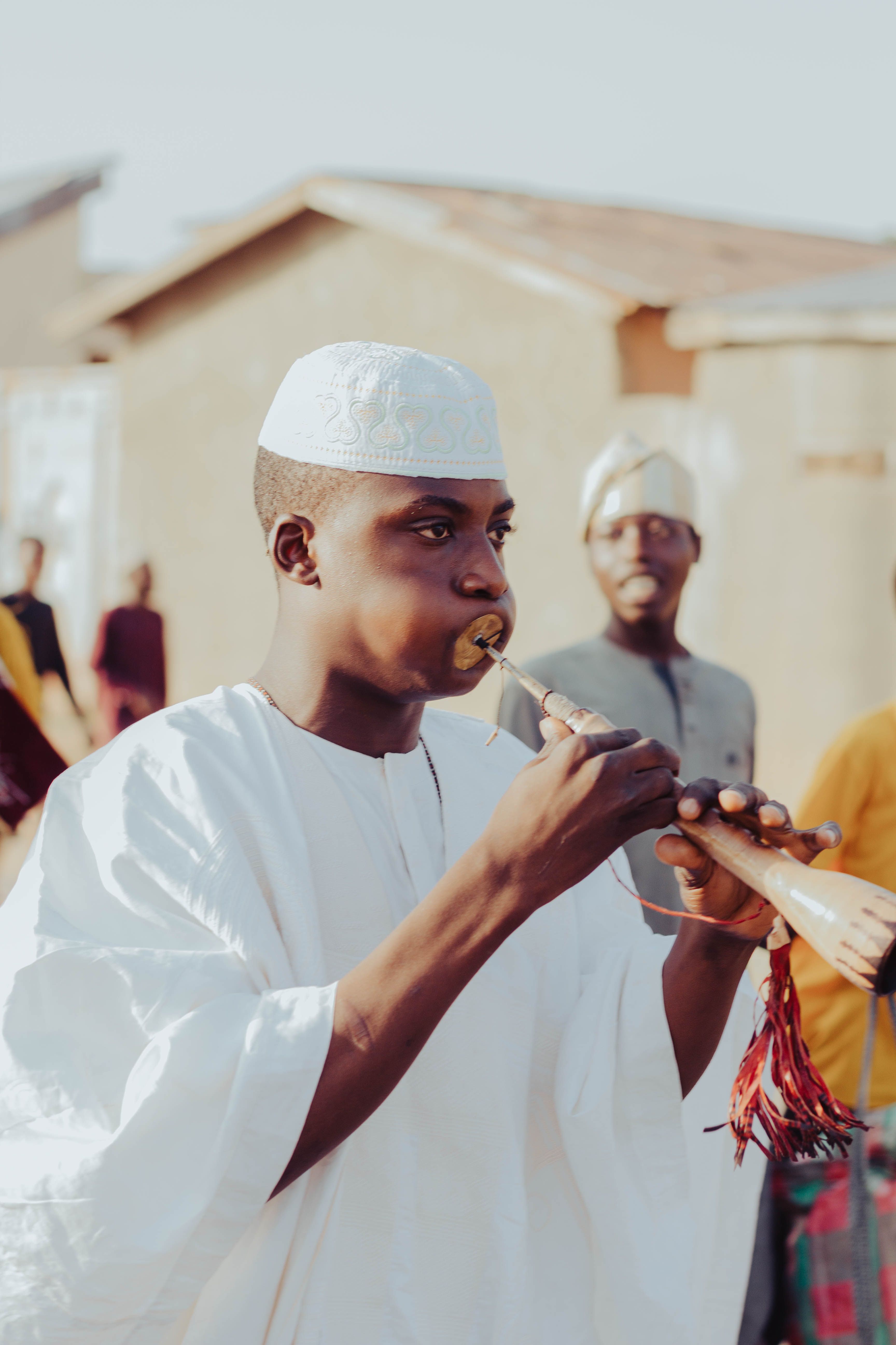 Man in traditional African attire playing a wooden flute outdoors during a cultural gathering.