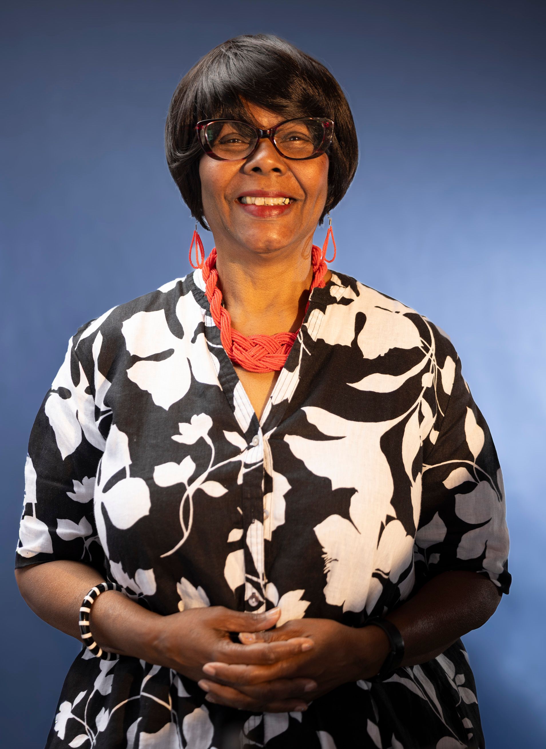 Elegant portrait of a woman wearing glasses, a patterned dress, and bold accessories against a blue backdrop.