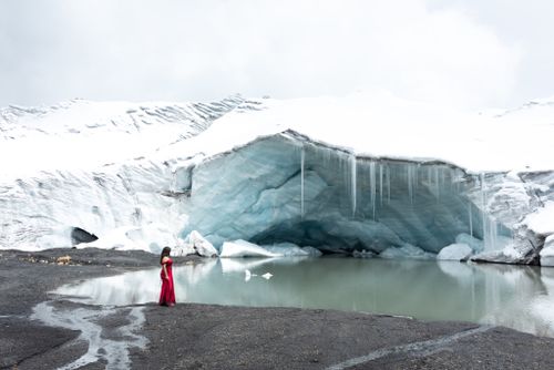 Free A woman in a red dress stands beside an ice cave with a serene water view. Stock Photo