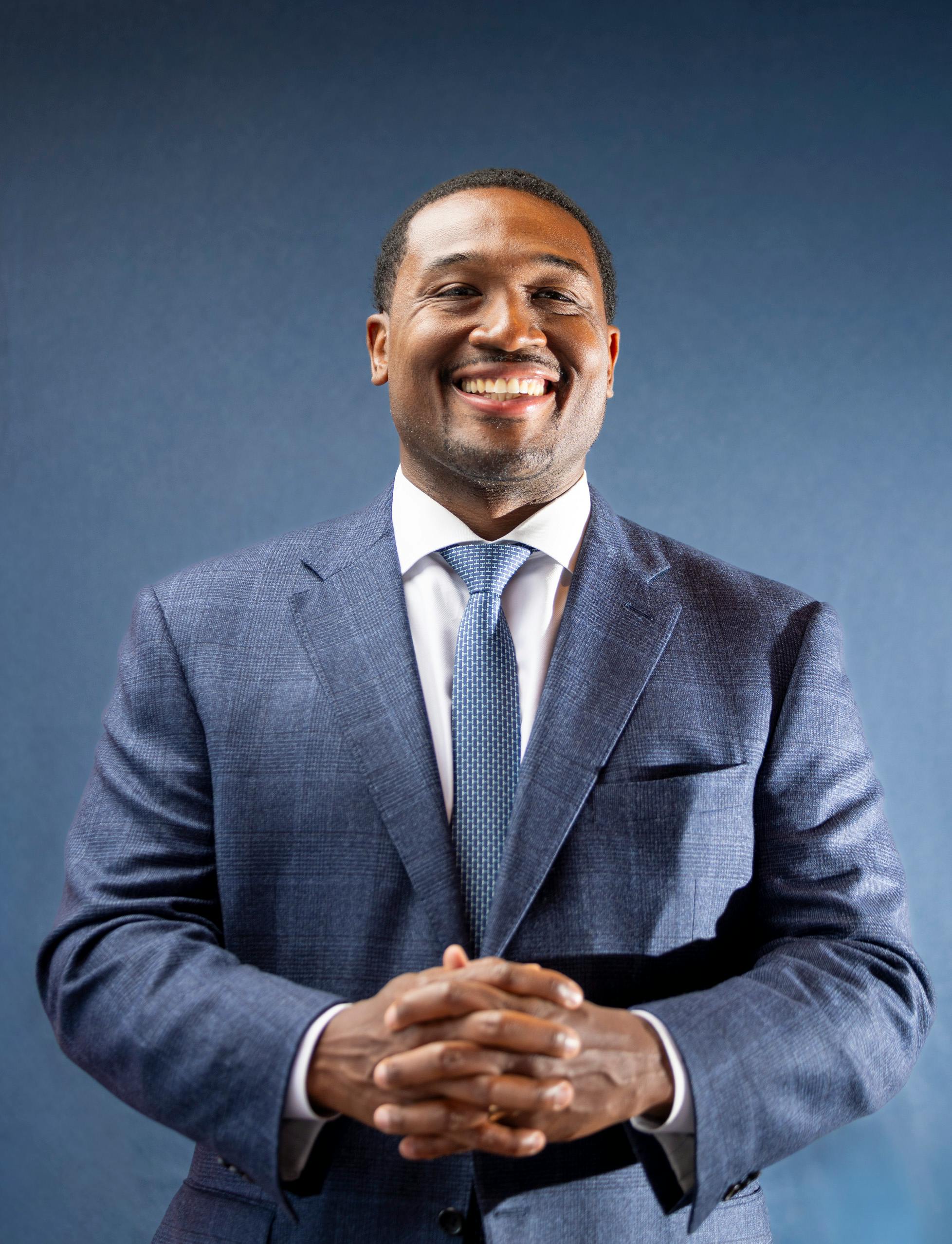 Smiling businessman in blue suit with confident posture standing against a dark background.