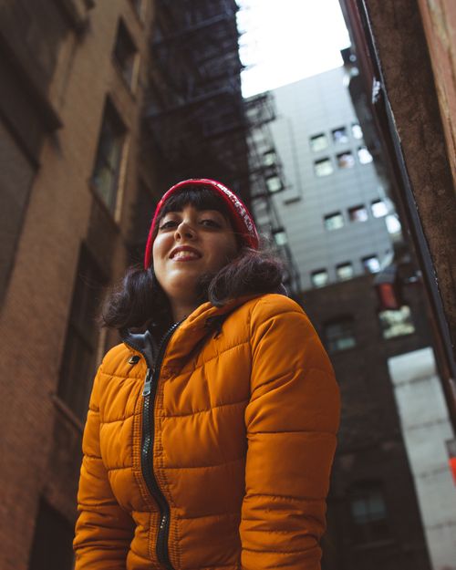 Free Woman in orange jacket and red beanie smiling in a city alleyway, showcasing winter fashion. Stock Photo
