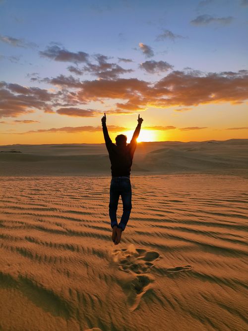 Free Person jumping joyfully in desert with sunset sky. Golden light creates dramatic scenery. Stock Photo
