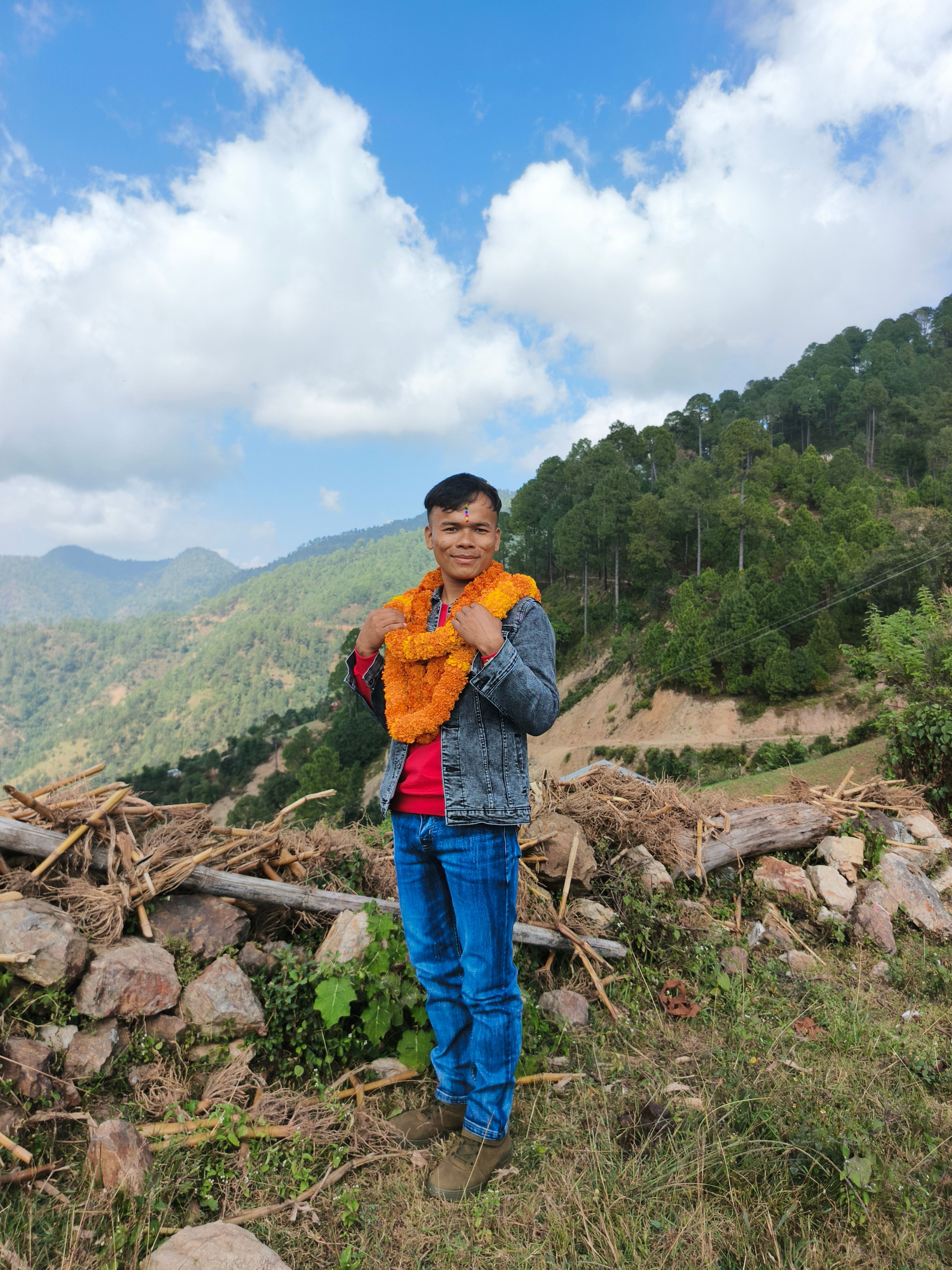 Man adorned with garland stands amidst lush hills and clear sky view.