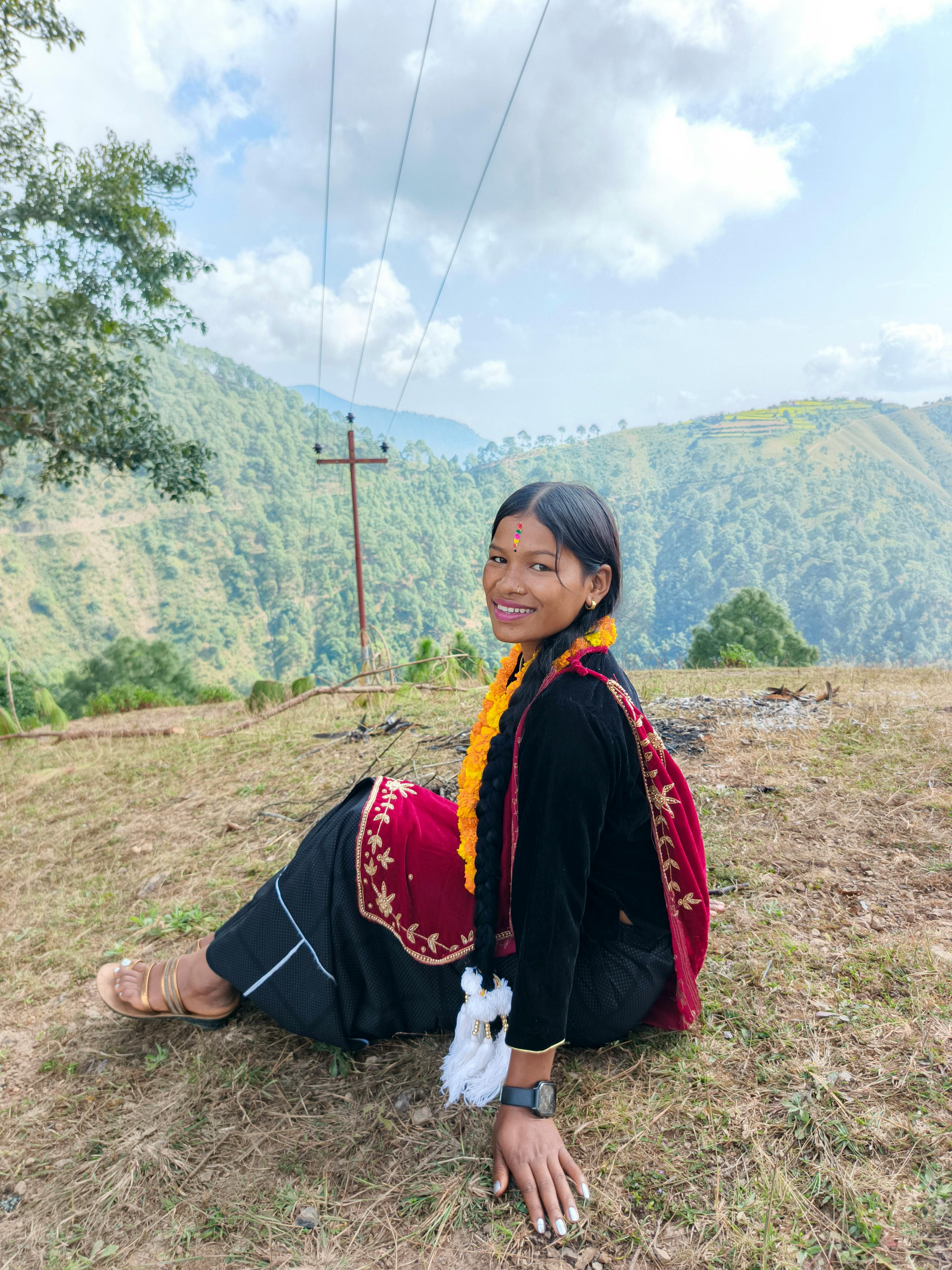Young woman in traditional attire sitting on a hill with a scenic mountain backdrop.