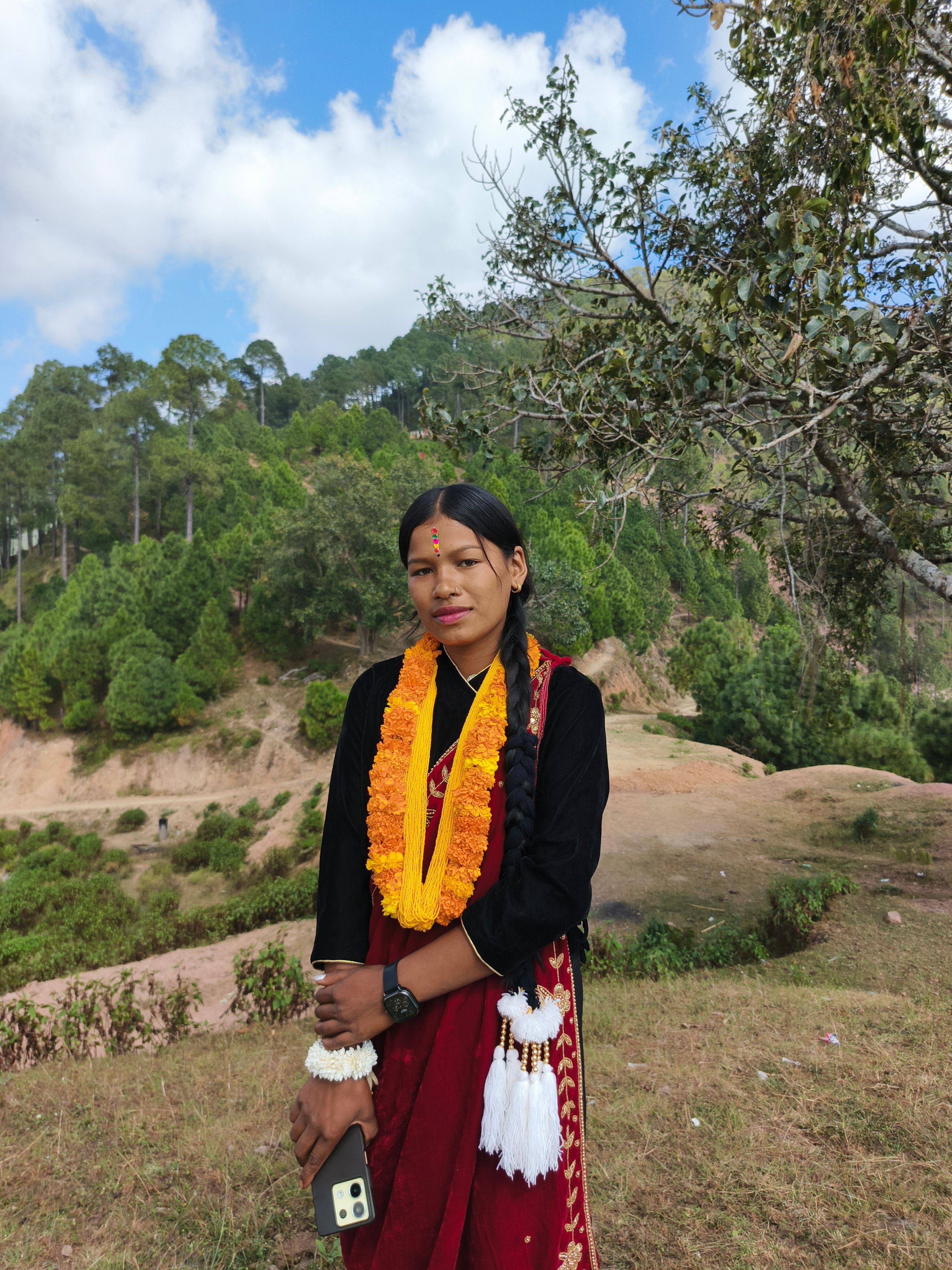 A woman in traditional clothing poses in a scenic outdoor setting with vibrant greenery.
