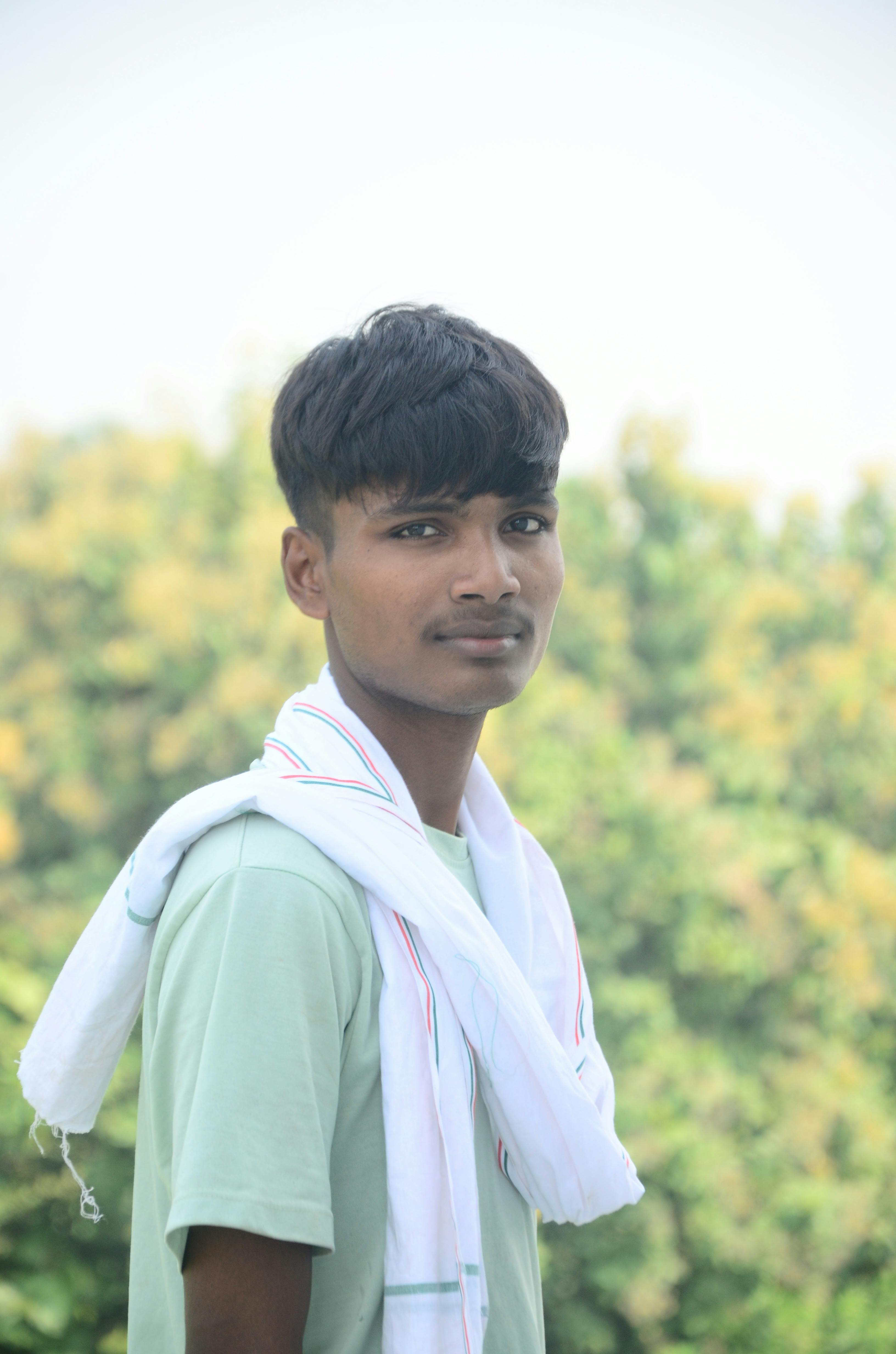 A young man stands outdoors with greenery in the background, wearing a casual shirt and a white scarf.