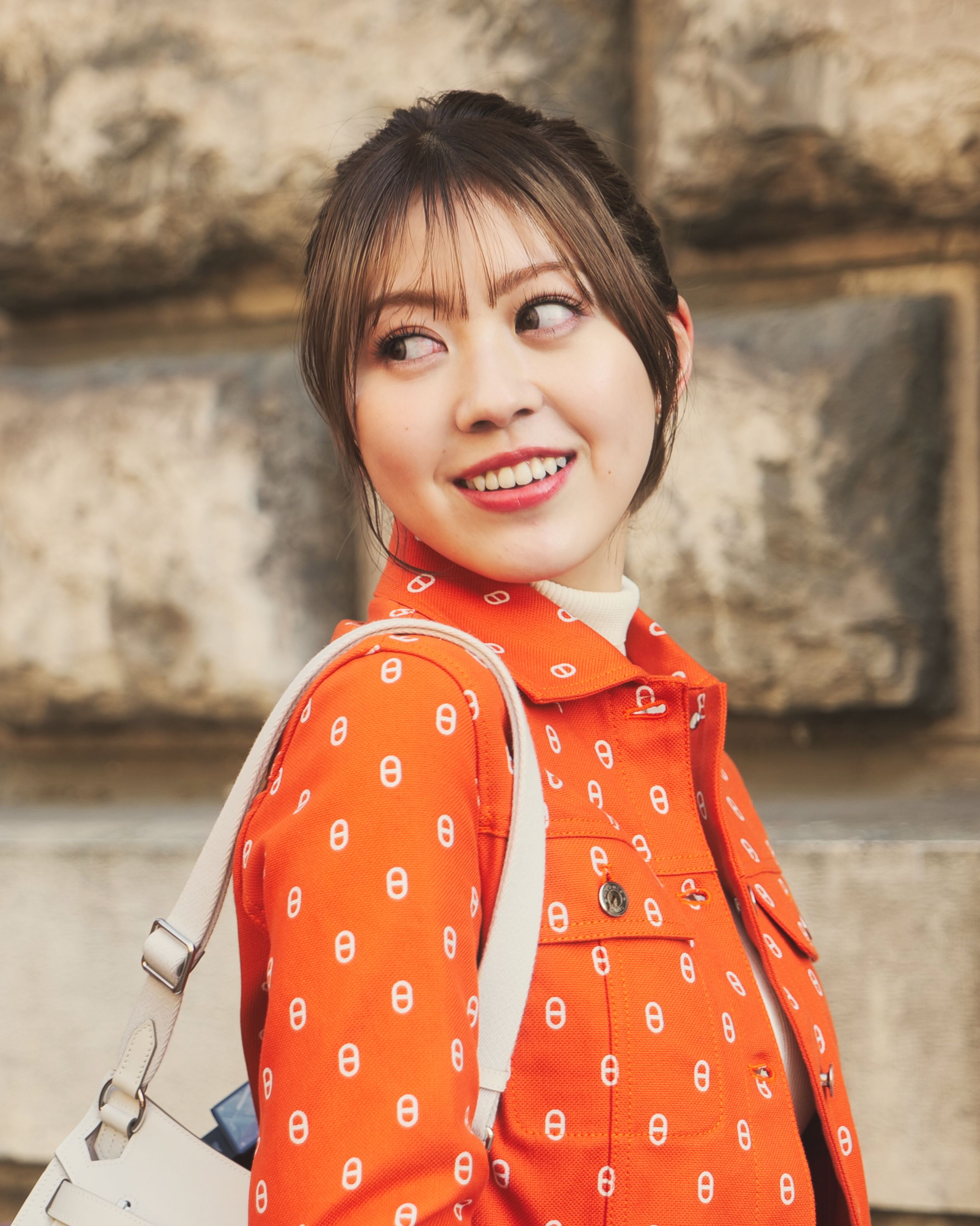 Elegant young woman in orange jacket smiling outdoors with natural background.