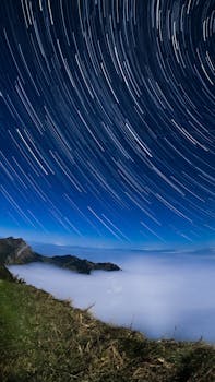 Dramatic star trails over clouds and alpine mountains under a clear night sky.