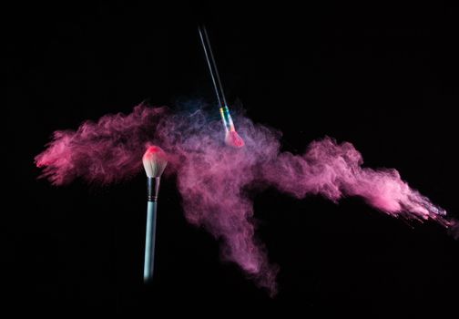 Two makeup brushes dynamically releasing pink powder against a black background.