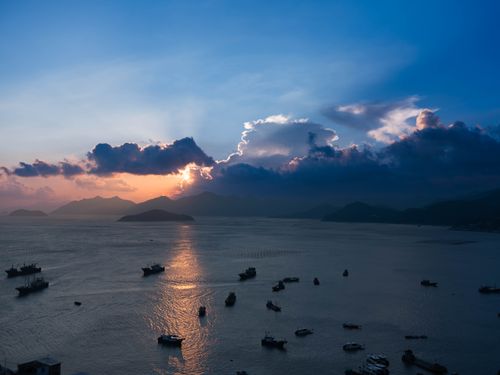 Free Serene sunset over a calm ocean with silhouetted boats and distant islands under dramatic clouds. Stock Photo