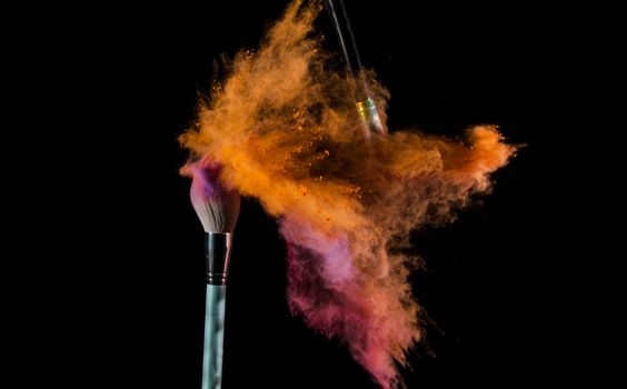 Dramatic splash of colorful powder with makeup brushes on black background.