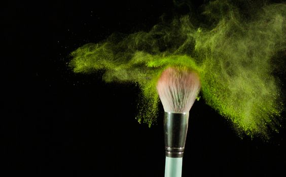 A makeup brush dispersing vibrant green powder against a black background.