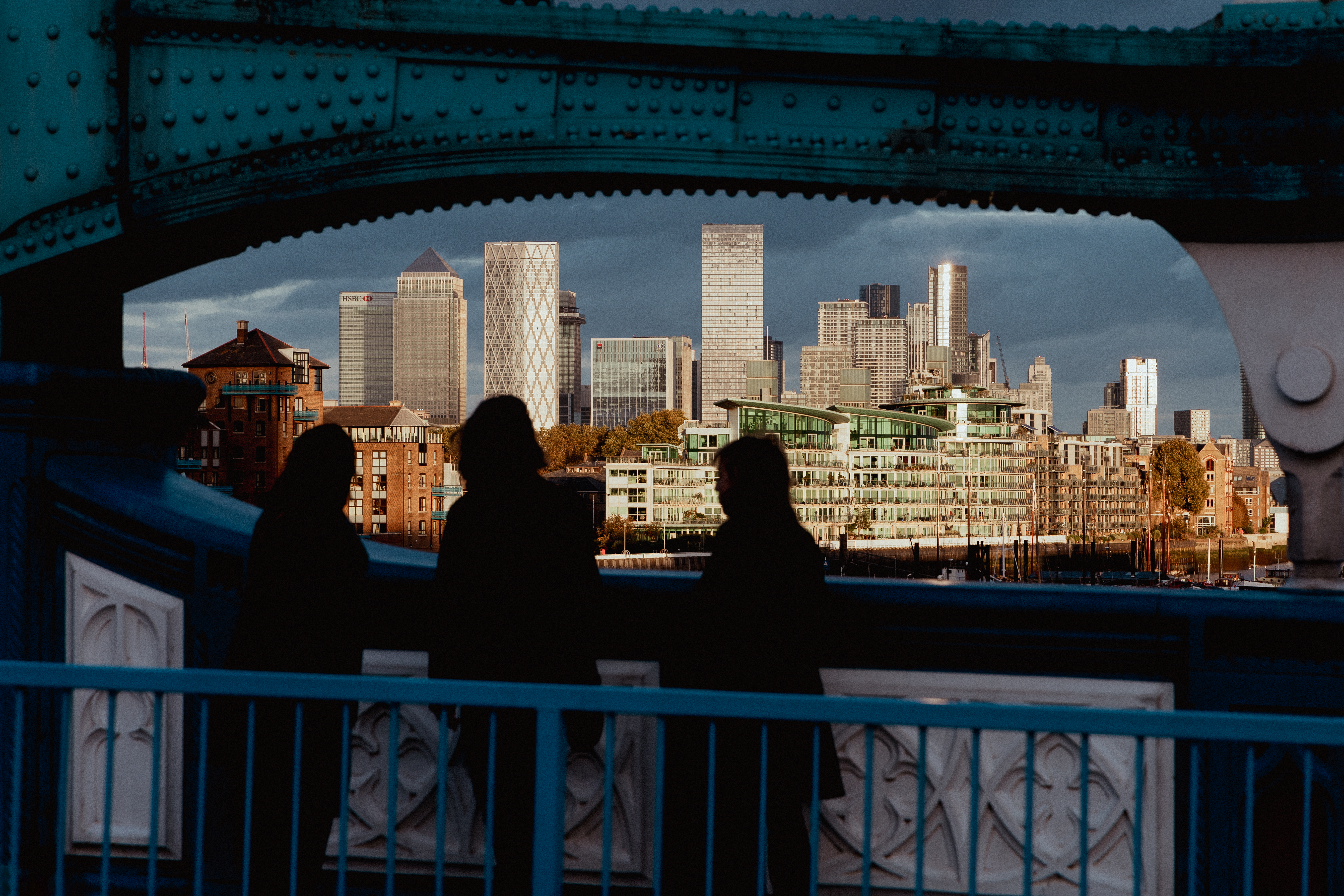 Three silhouetted figures frame a view of Canary Wharf's modern skyline at sunset from a London bridge.