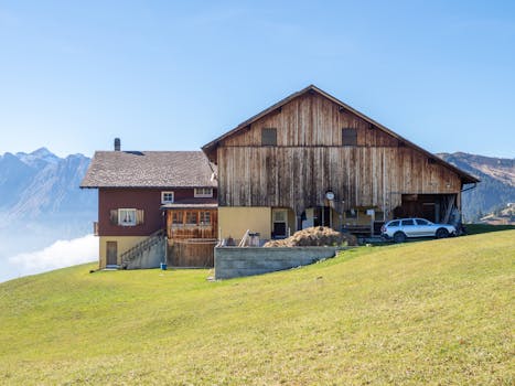 A rustic farmhouse with wooden siding nestled in a mountainous landscape under a clear blue sky.
