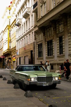 A classic green Cadillac parked on a sunlit European street with pedestrians walking by.