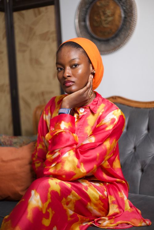 Free Elegant woman in a vibrant tie-dye outfit with an orange headwrap, sitting indoors on a sofa. Stock Photo