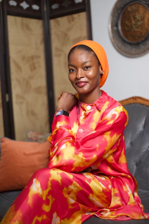 Free Confident woman in colorful tie-dye dress with an orange headwrap, seated indoors. Stock Photo
