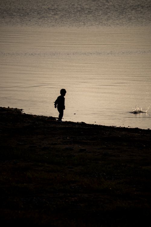 Free A child stands by the lakeside at dusk, silhouetted against the calm water. Stock Photo
