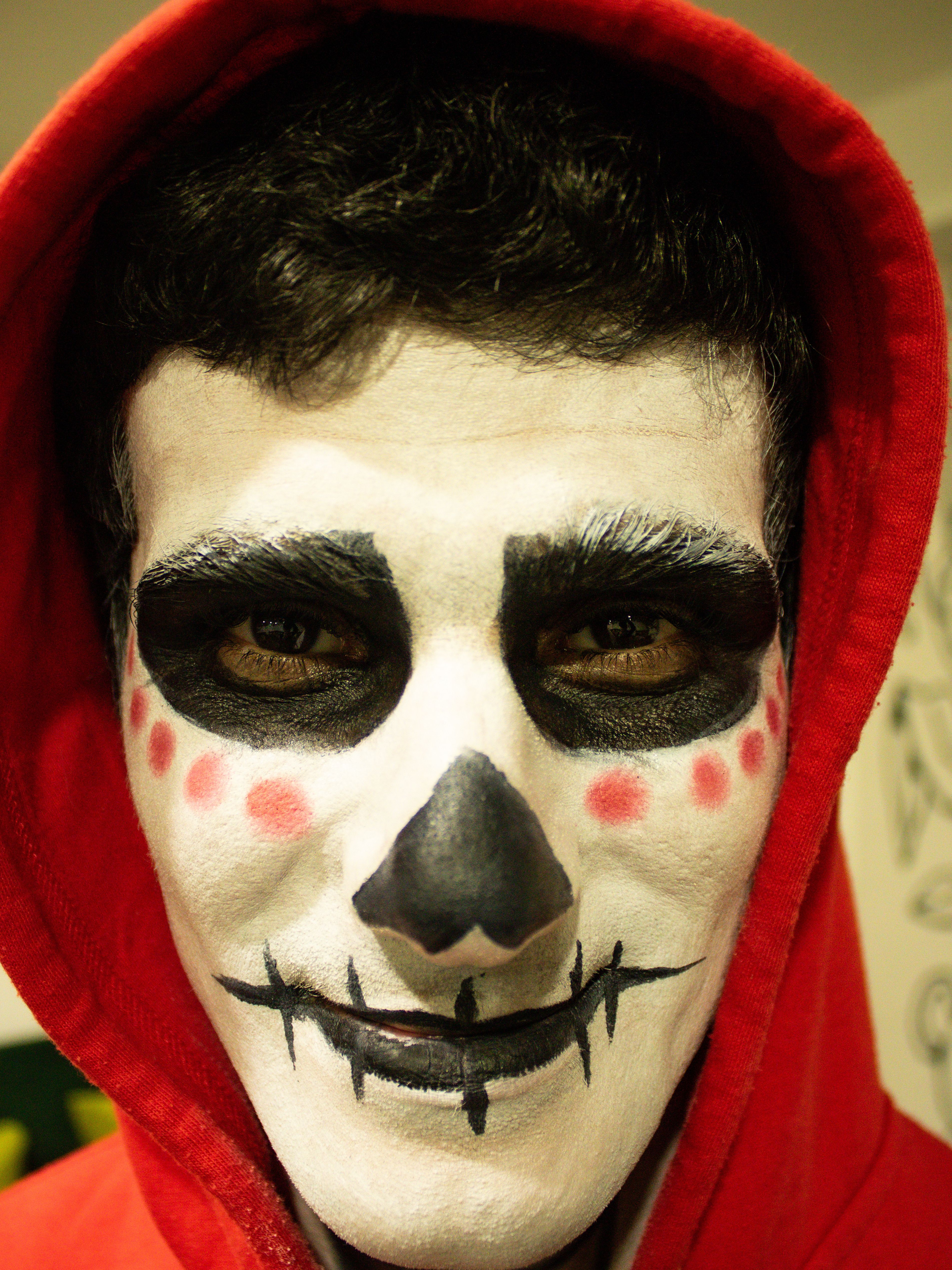 Person with intricate skull face paint wearing a red hood, perfect for Halloween or Day of the Dead.