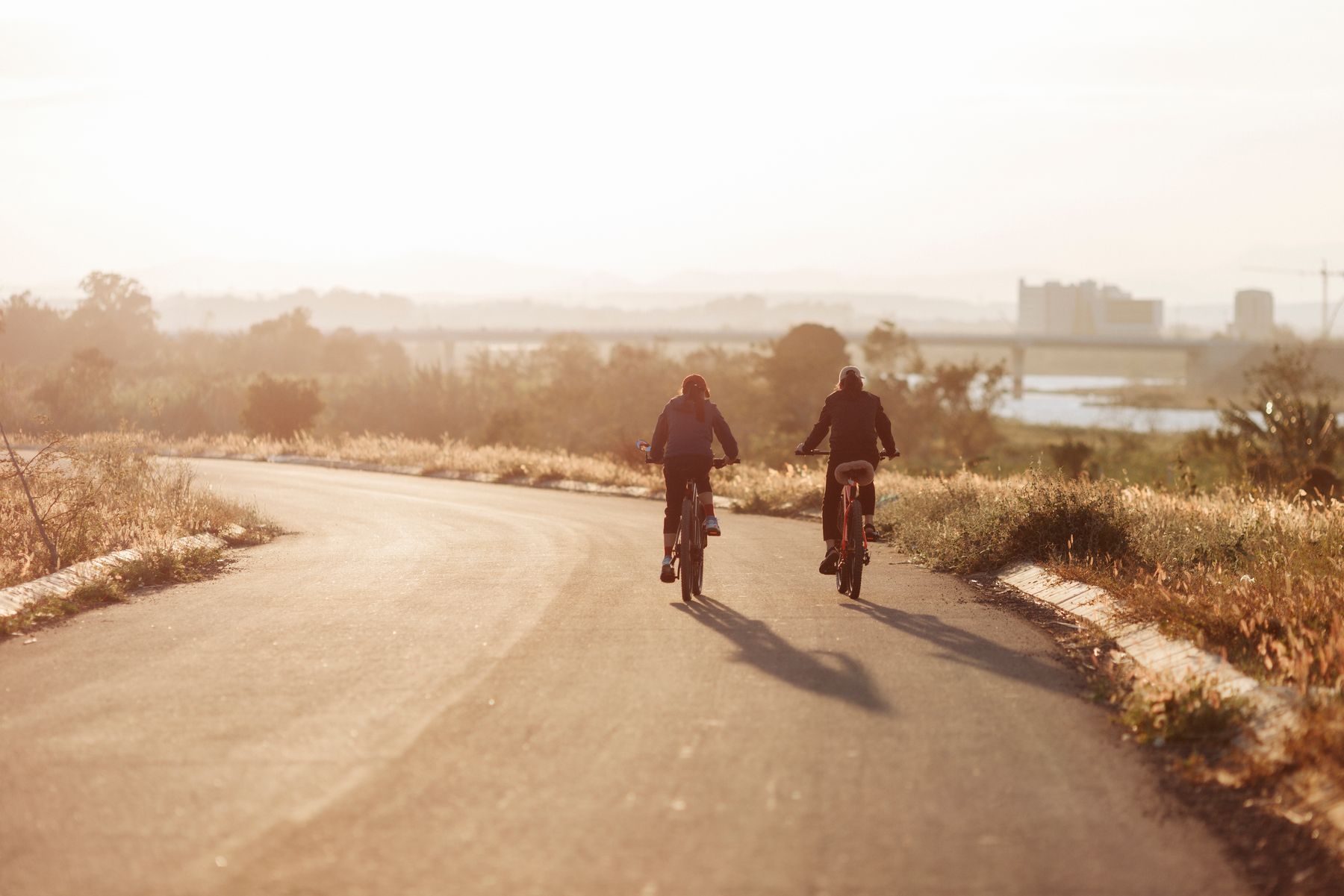 Two cyclists riding side by side along a curving country road in hazy golden evening light, long shadows trailing behind them