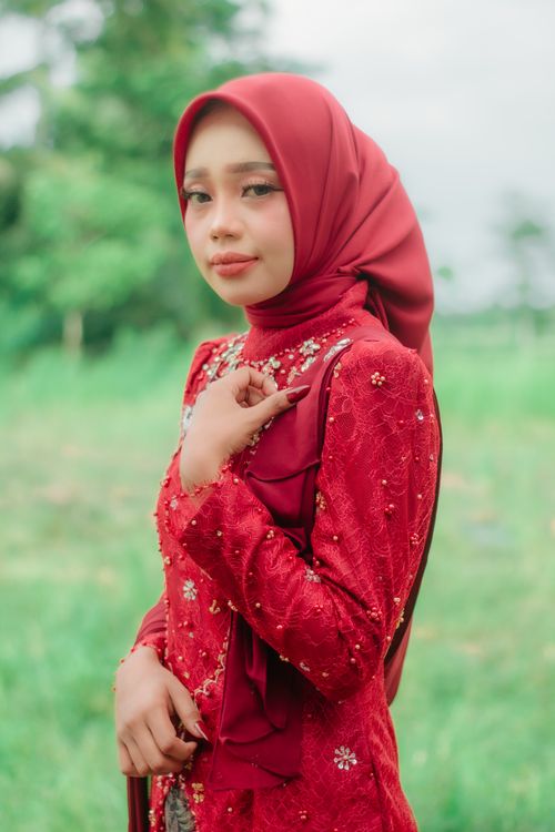 Free Portrait of a woman wearing a red hijab and dress, posing outdoors with a blurred green background. Stock Photo