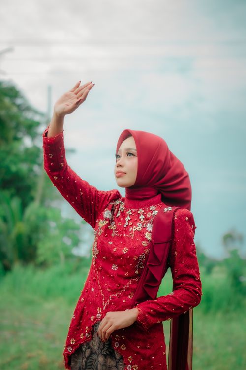 Free Woman in a red floral dress and hijab outdoors, with hand raised gracefully. Stock Photo