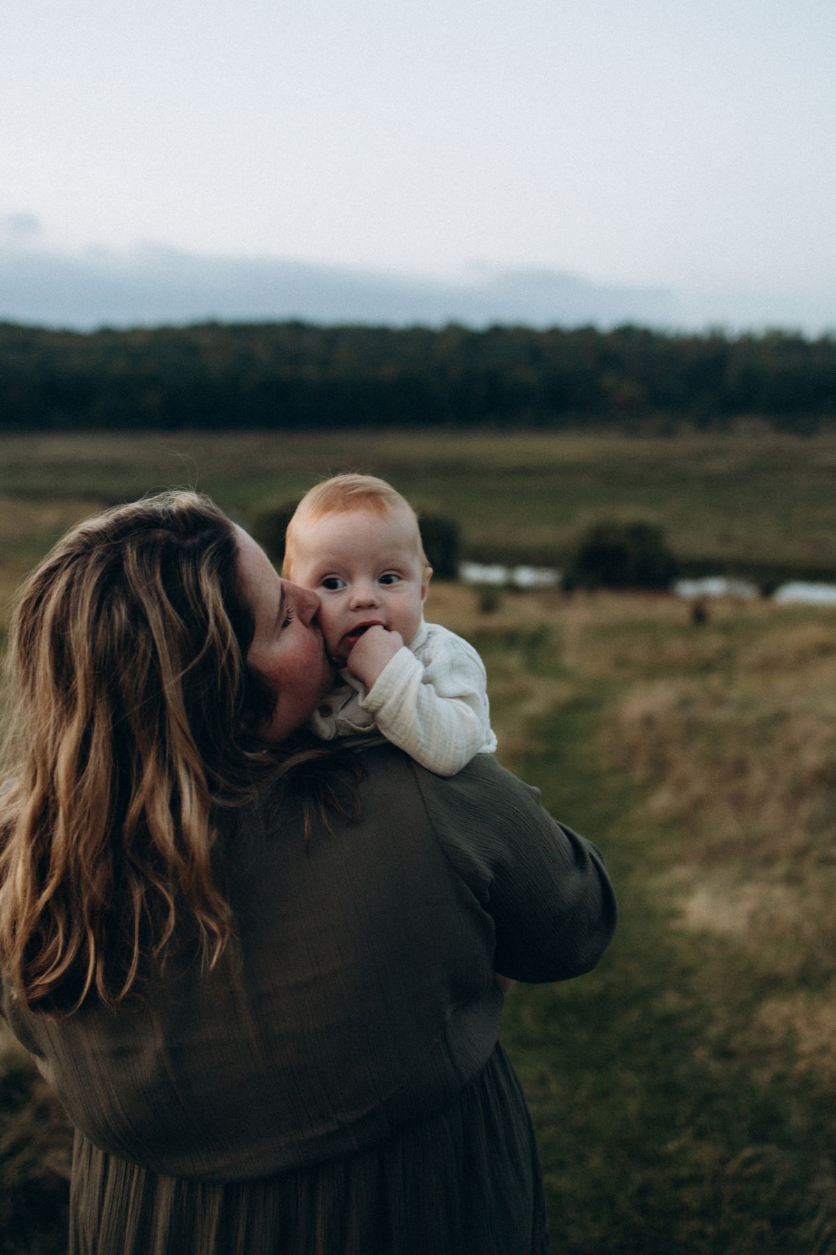 A mother kissing her baby's cheek as she carries them over her shoulder across a grassy field on an overcast evening