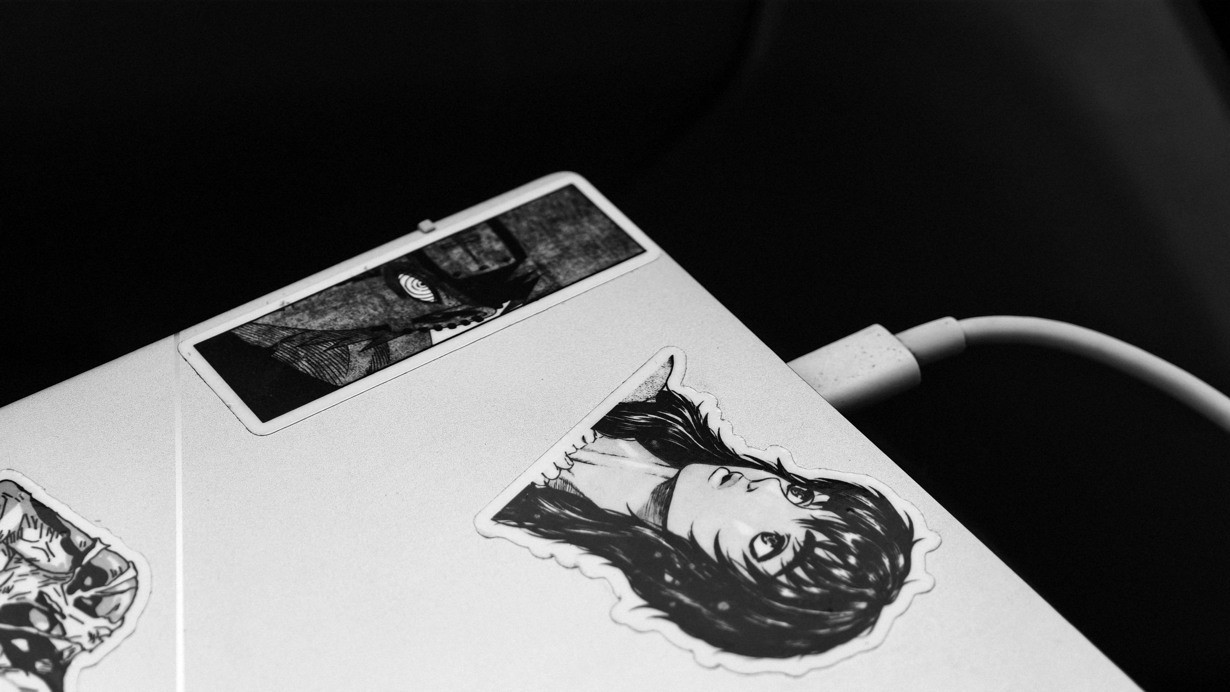 Gratis Gambar monokrom laptop yang dihiasi stiker bertema anime dan terhubung ke pengisi daya. Foto Stok
