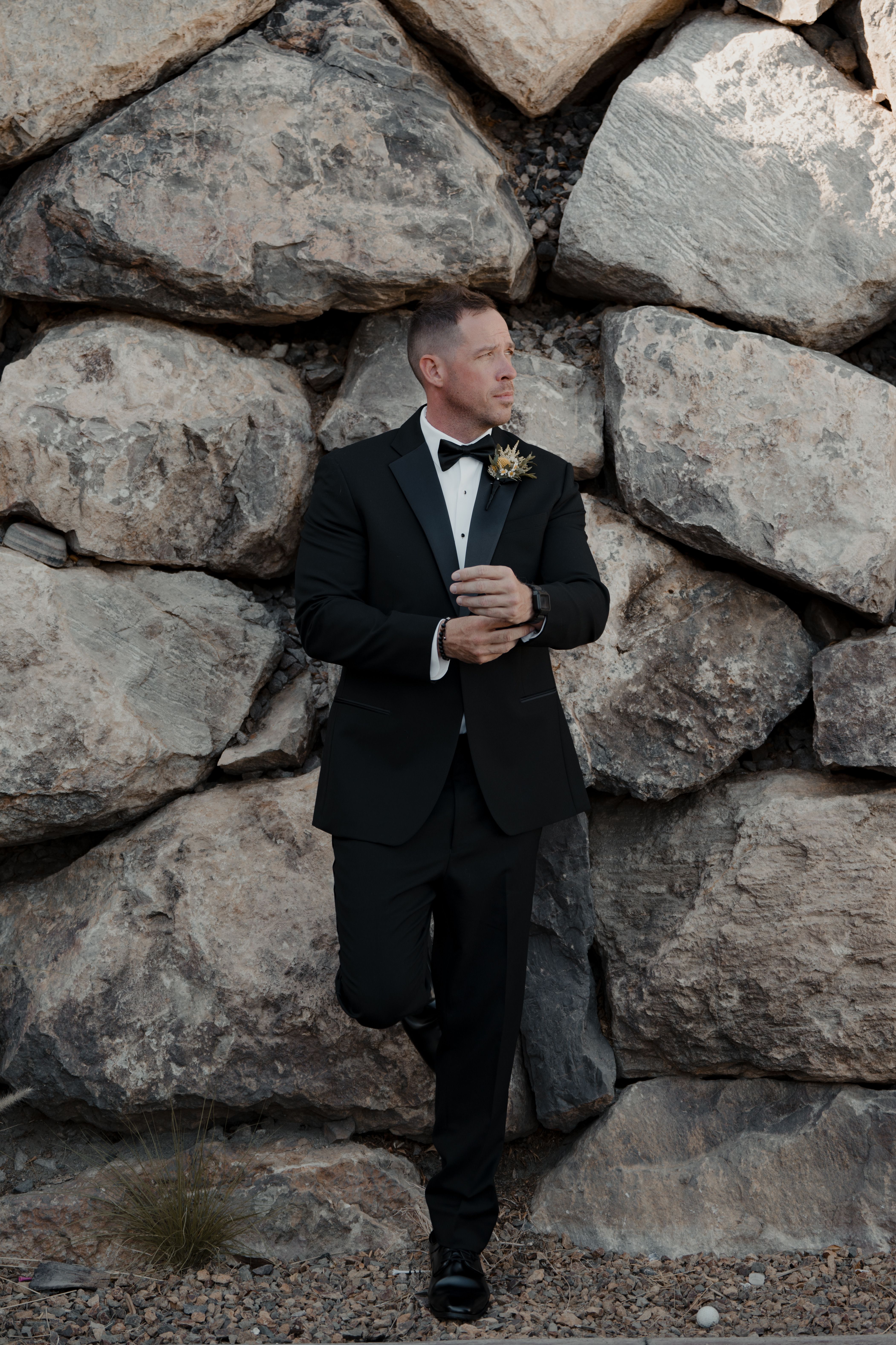 Man in formal wear standing by a rustic rock wall, exuding elegance and sophistication.