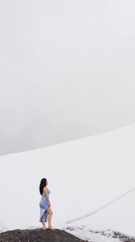 A woman in a blue dress stands on a snowy mountain, creating a striking contrast.