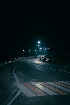 Eerie night view of a winding road lit by streetlights, featuring a pedestrian crossing.
