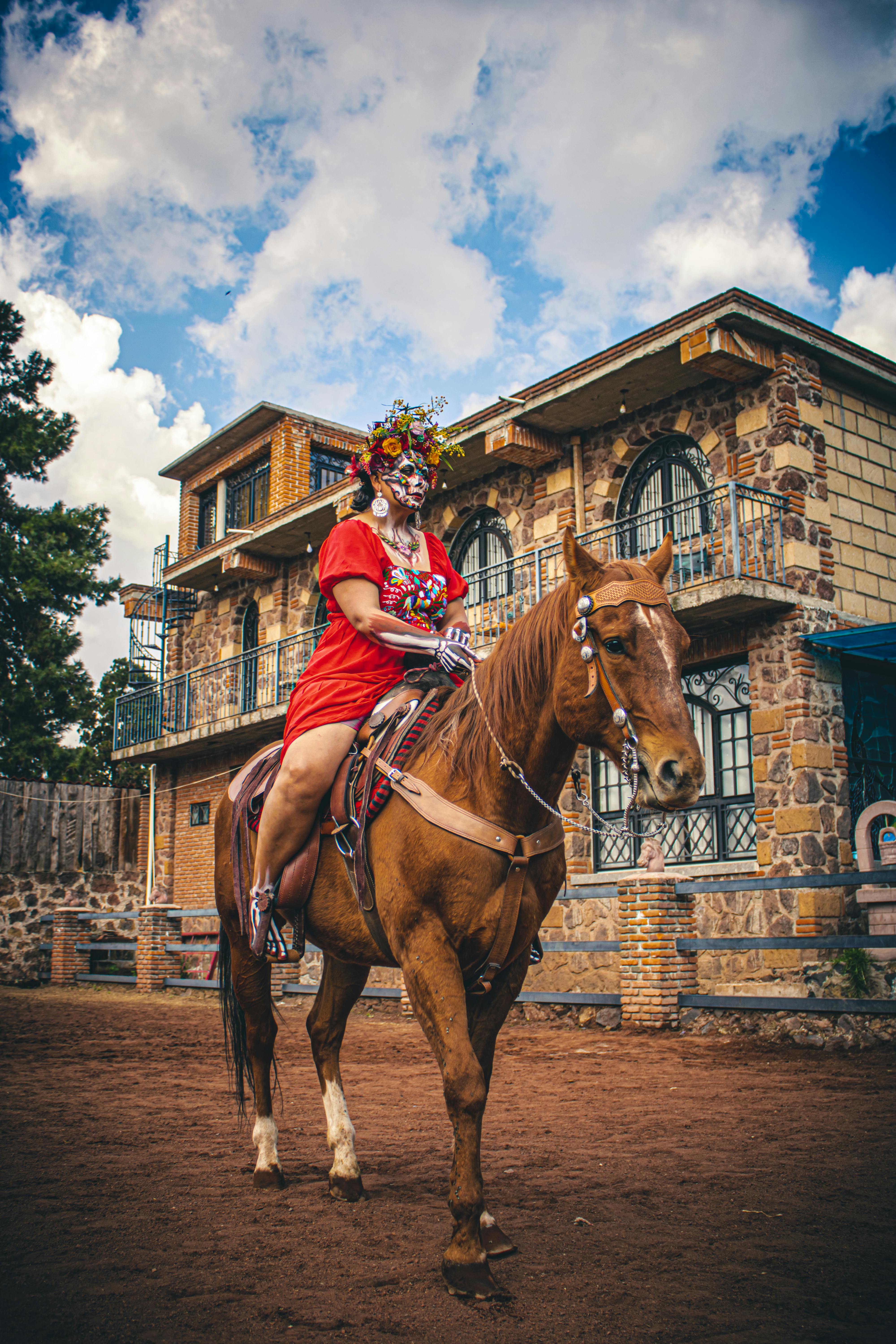 A woman in Catrina makeup riding a chestnut horse in front of a traditional Mexican building.