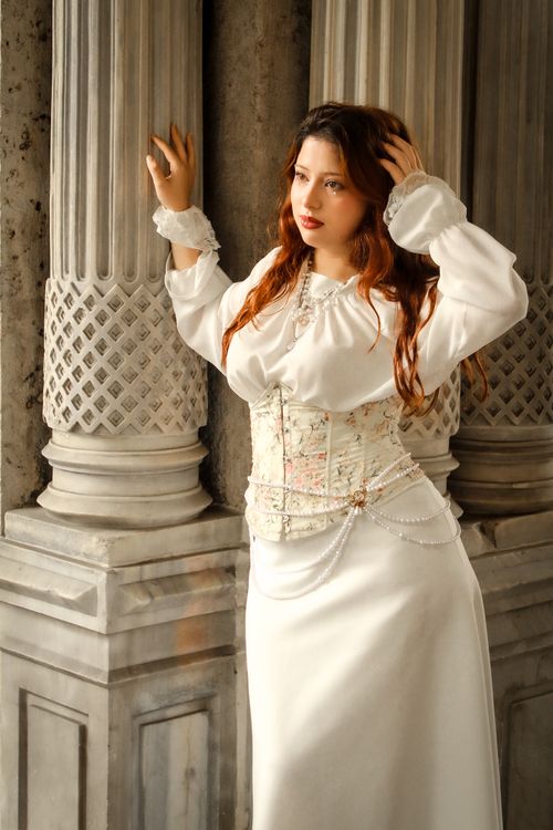 Free Woman in a white Victorian-style dress standing by ornate columns, exuding elegance. Stock Photo