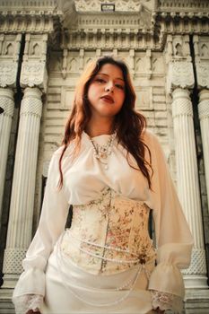 Stylish woman in Victorian-style attire poses against intricate column architecture, exuding elegance and grace.