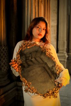 A woman in a white dress holds a decorative mirror amid classical architecture, lit warmly by sunset.