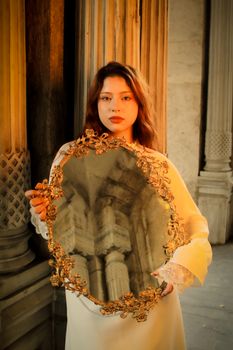 A young woman holding a decorative mirror in front of elegant architectural columns.