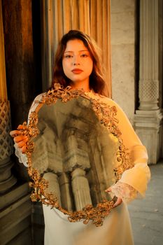 Elegant woman in a white dress holding a decorative mirror reflecting historical columns.