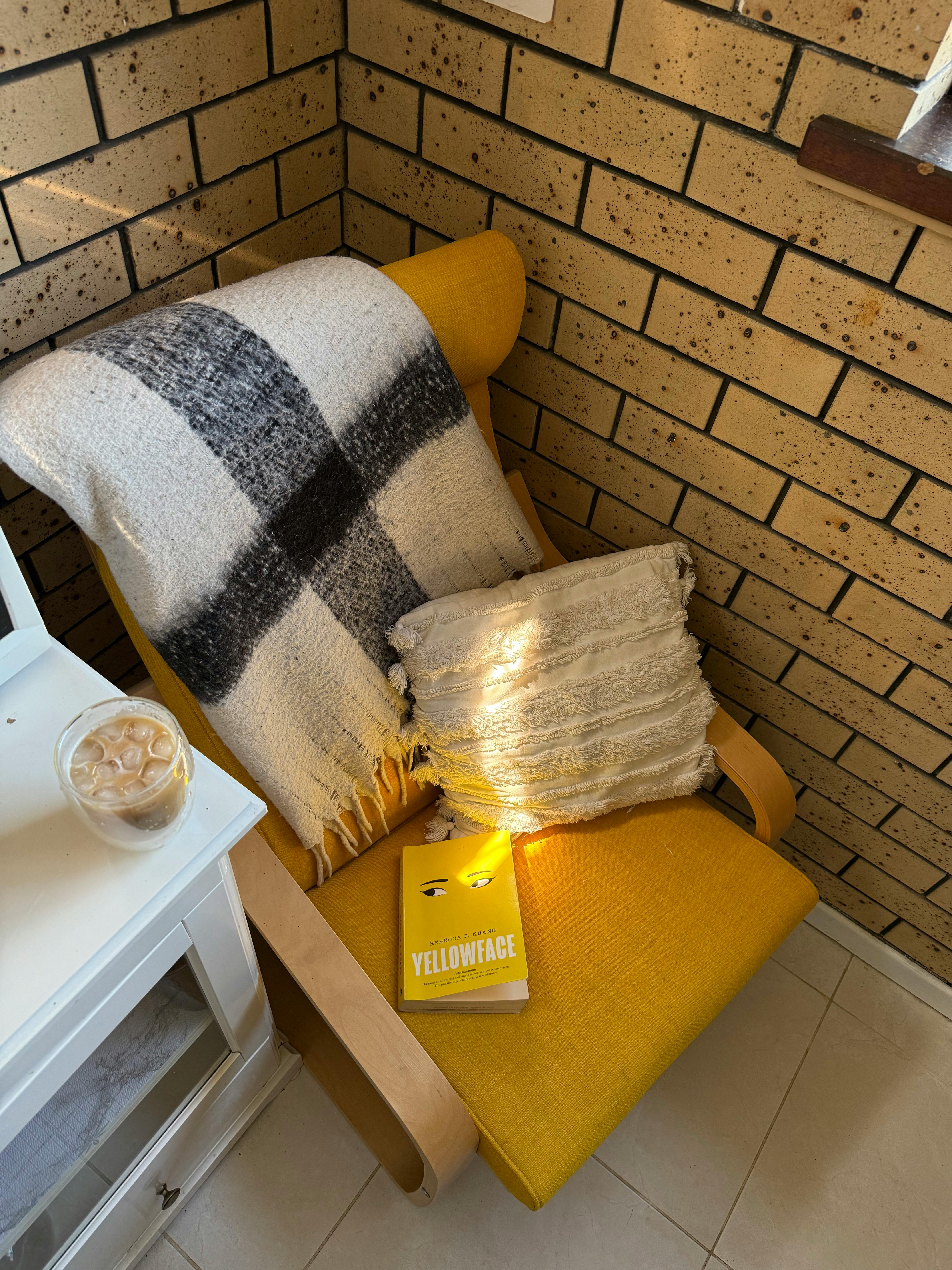 Bright corner armchair with blanket, pillow, book 'Yellowface', and iced coffee.