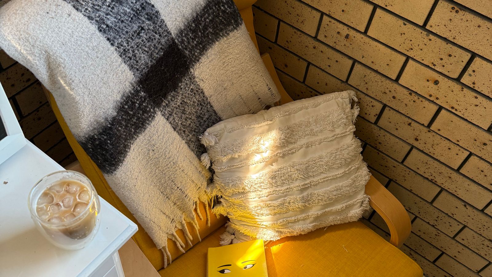 A yellow chair with a blanket, cushion and book beside a table