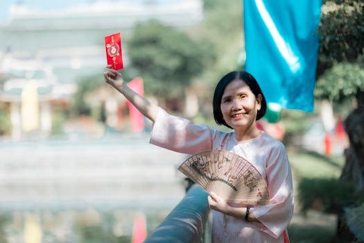 Smiling woman in traditional attire holding a red envelope and fan outdoors, symbolizing celebration.