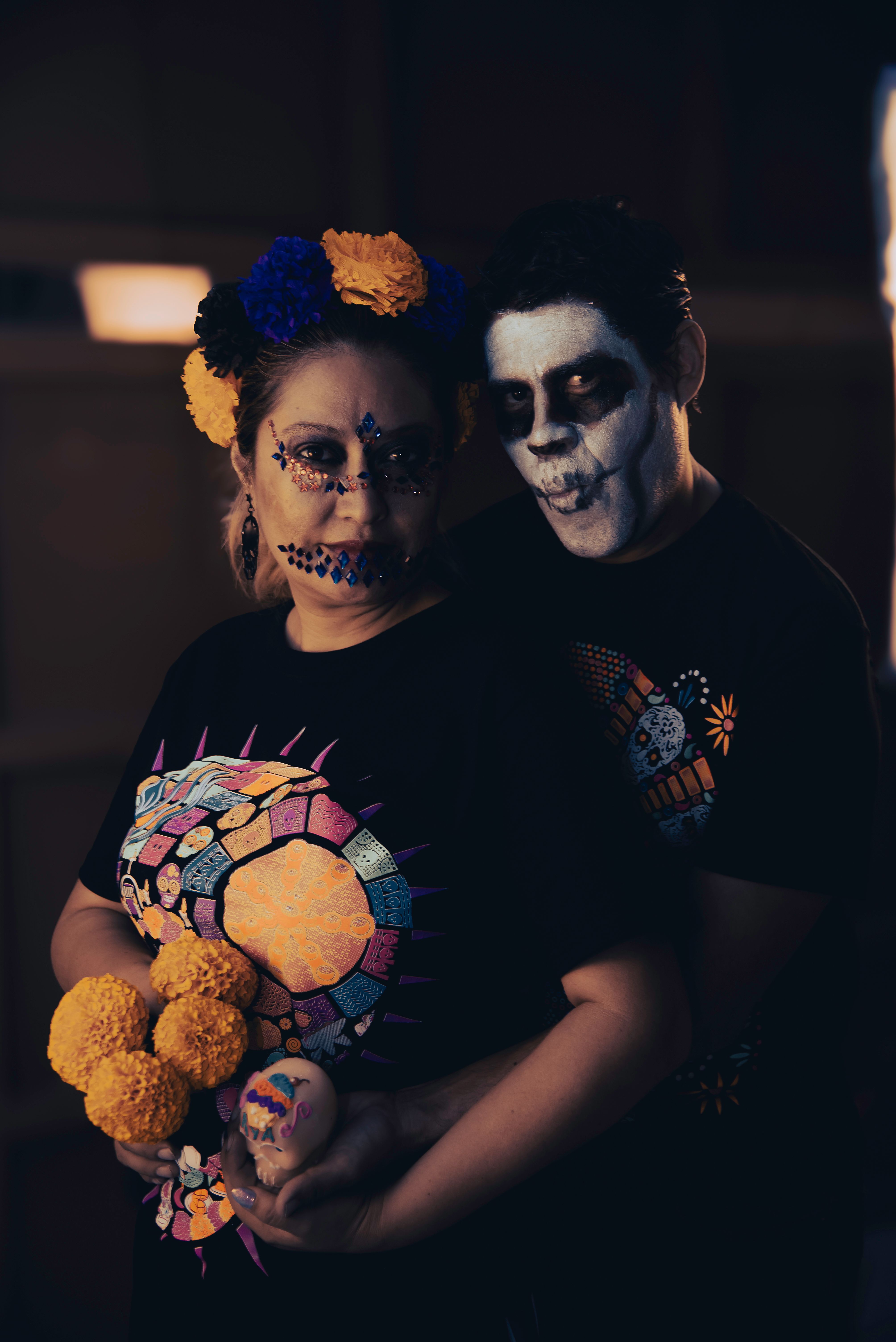 A couple in traditional Día de los Muertos makeup holding marigolds and decorated eggs.