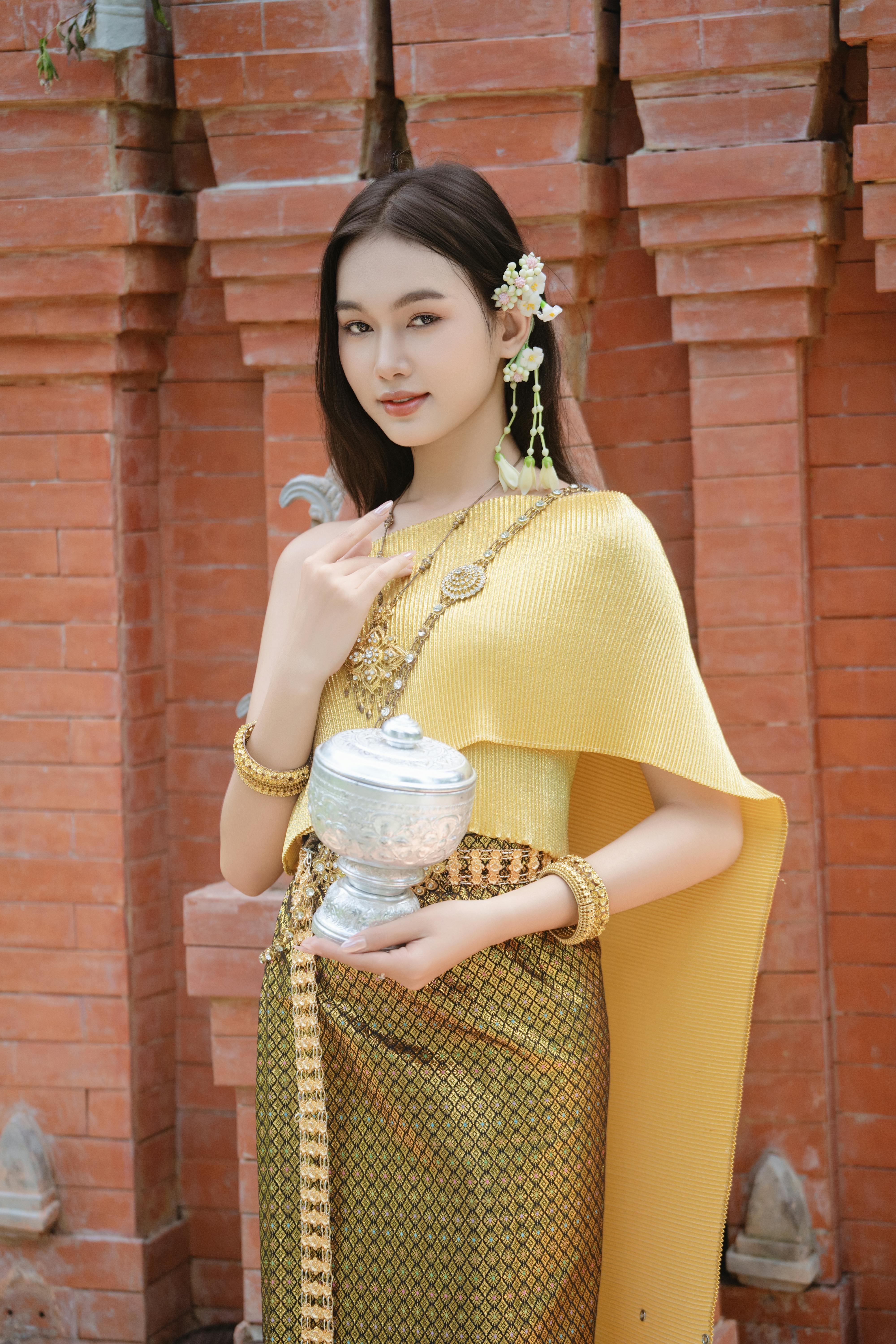 Elegant woman in traditional Thai dress holding a silver bowl, standing by a brick wall.