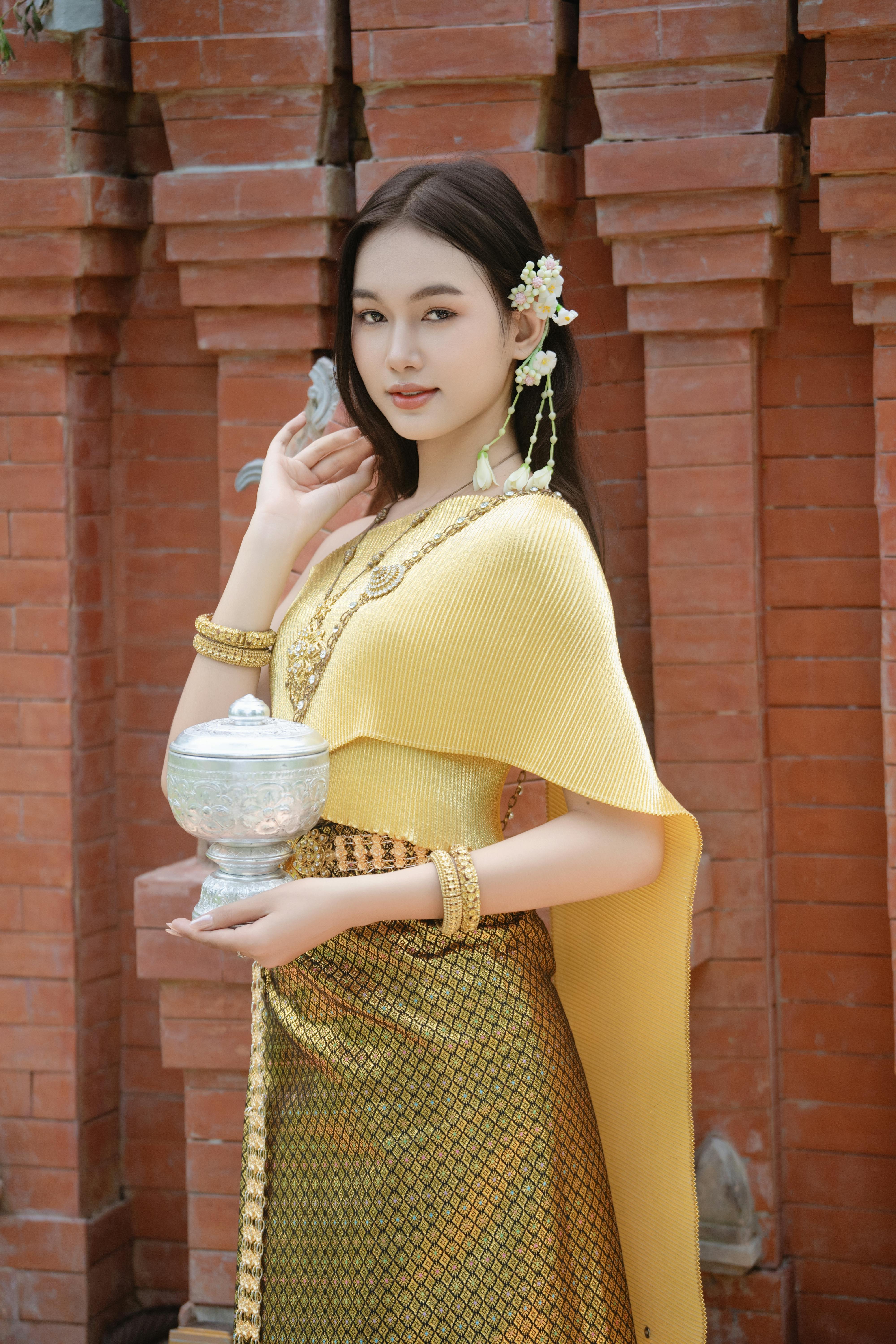 A woman in traditional attire holding a decorative silver bowl in front of a brick wall.