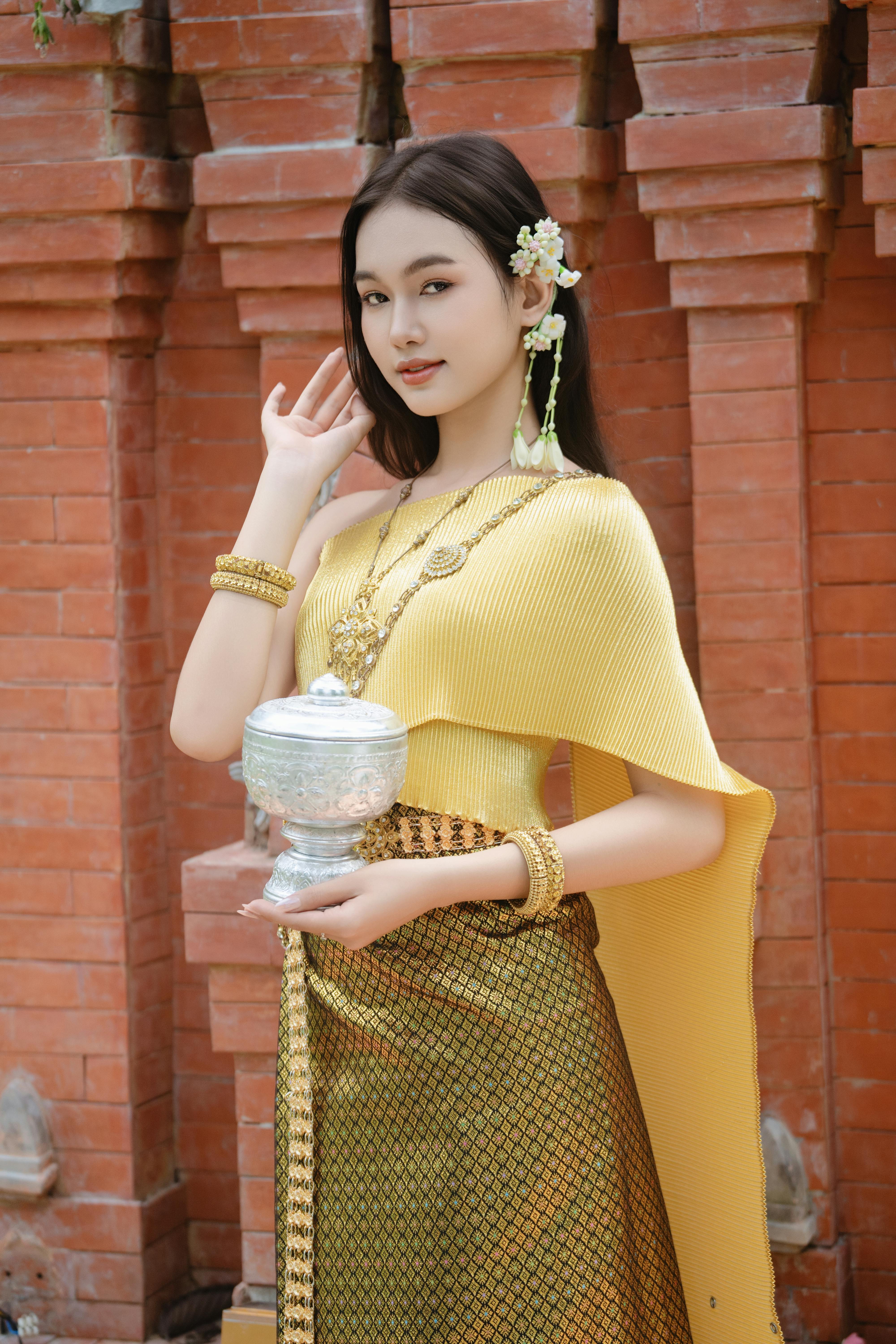 Young woman in traditional Thai attire holding a silver urn against a red brick wall.