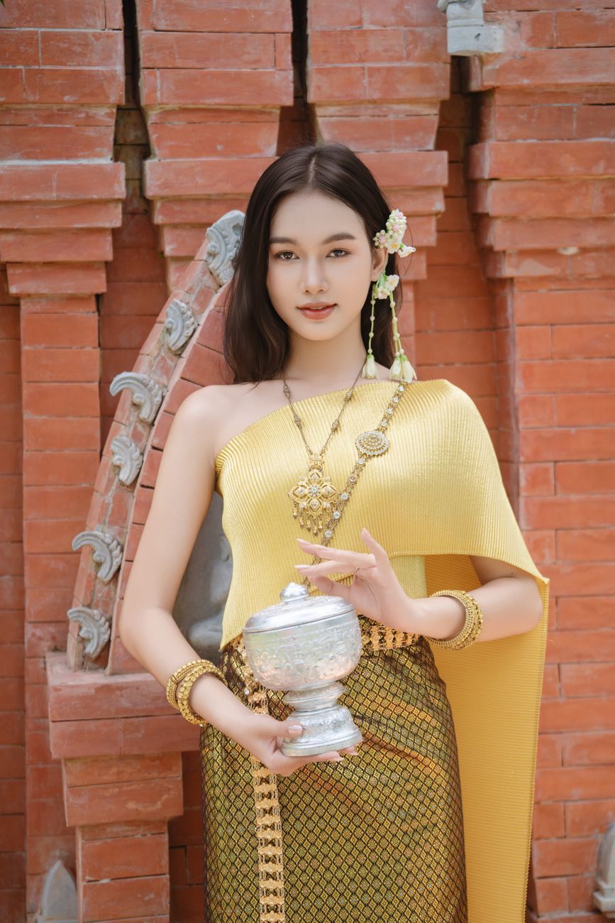 Elegant woman in traditional Thai attire holding a silver vessel, posing by a vintage brick wall.