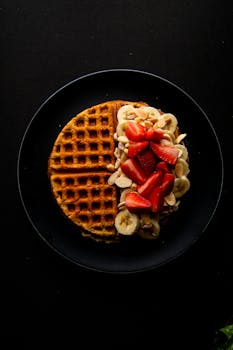 Savor this stunning waffle topped with fresh strawberries and bananas on a black plate.