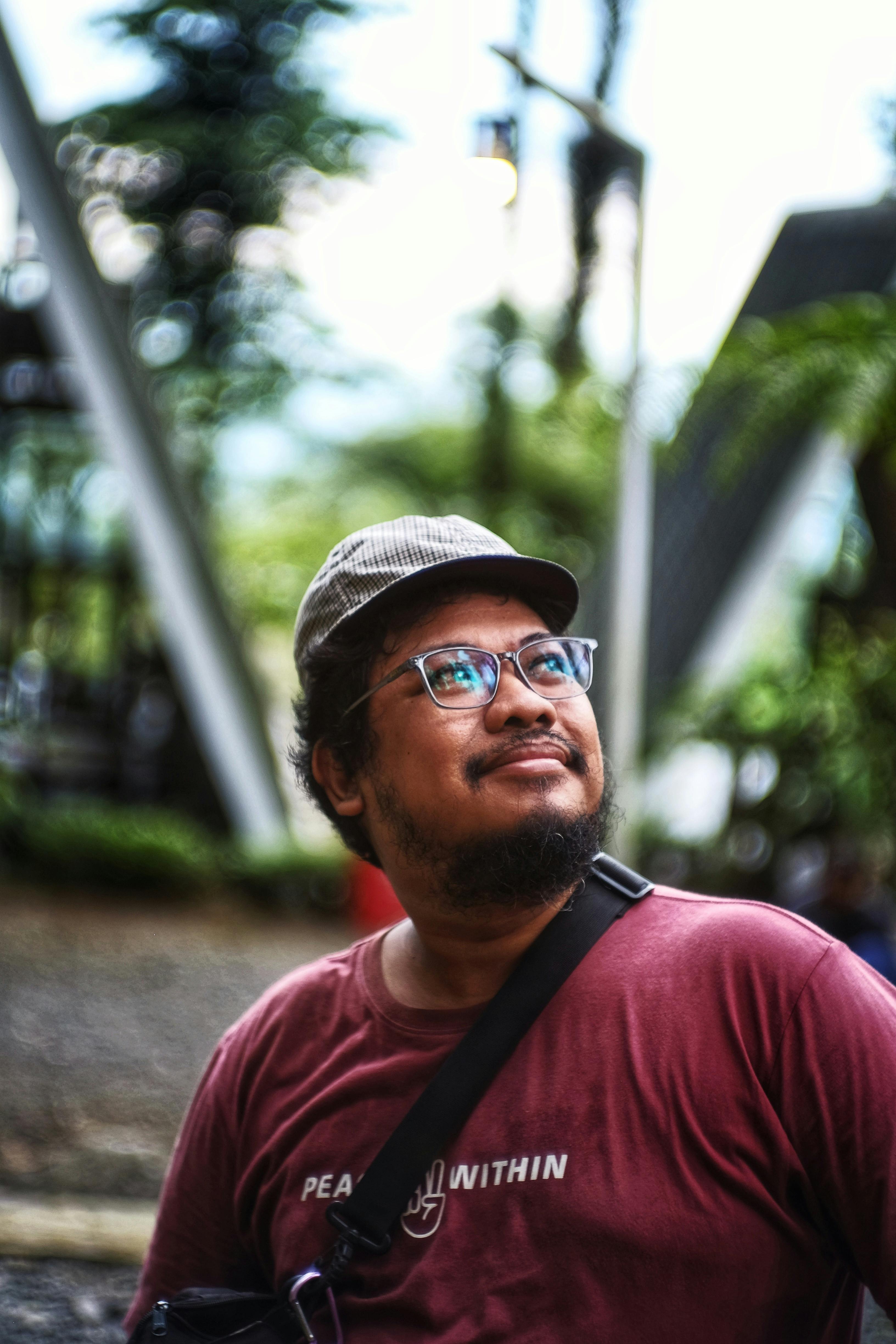 A thoughtful man outdoors, wearing glasses and a classic hat, gazing upwards.