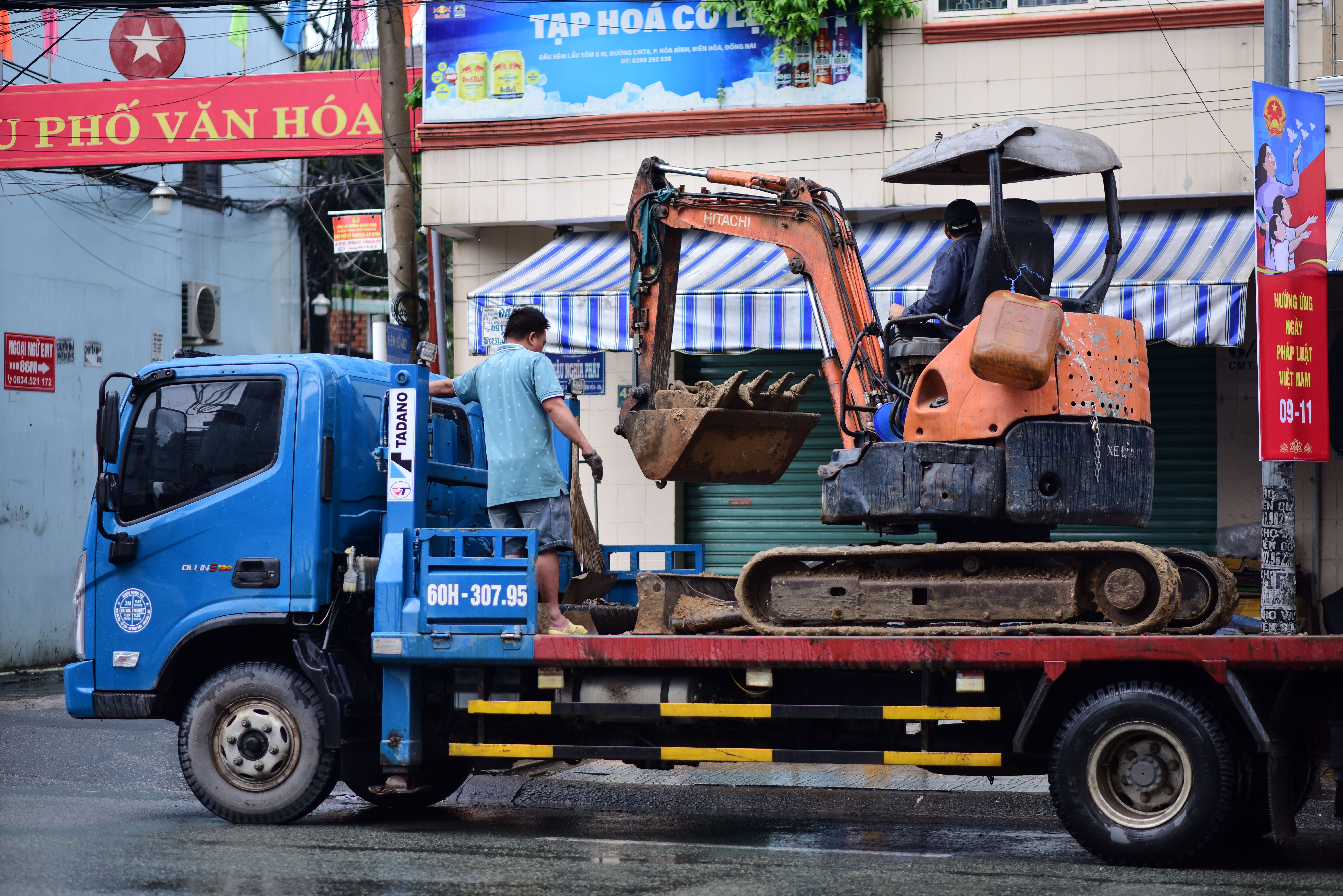 A blue flatbed truck carrying a small orange excavator on a city street with banners in the background.