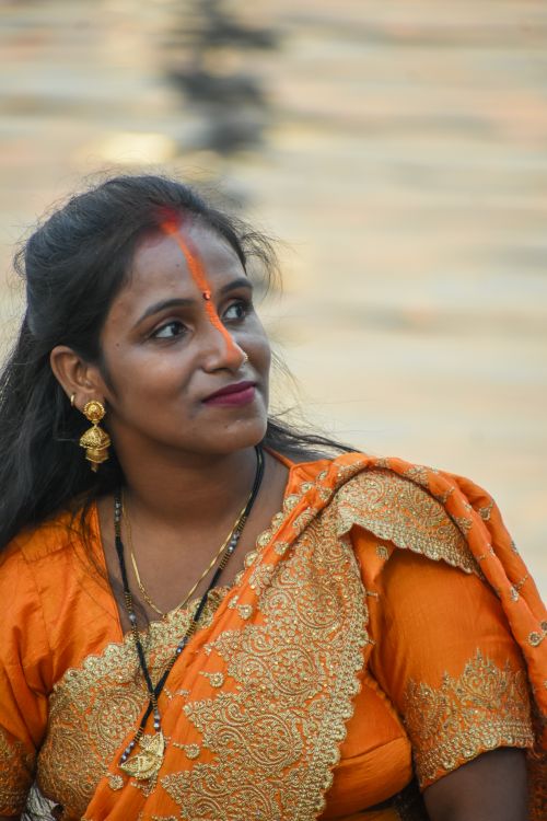 Free Portrait of a woman in an orange sari with traditional jewelry by the water. Stock Photo